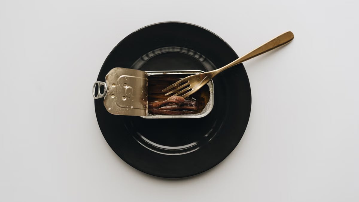 A can of anchovies served on a black plate with a fork in a minimalist setting.